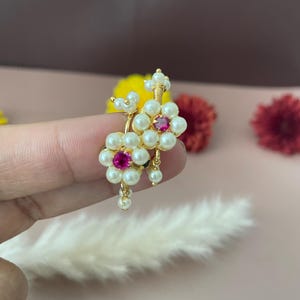 Pode incluir: Brincos florais dourados com detalhes de p&eacute;rolas brancas e uma gema rosa central. Os brincos t&ecirc;m um design delicado com pequenas p&eacute;rolas penduradas. O fundo &eacute; rosa suave com flores desfocadas.