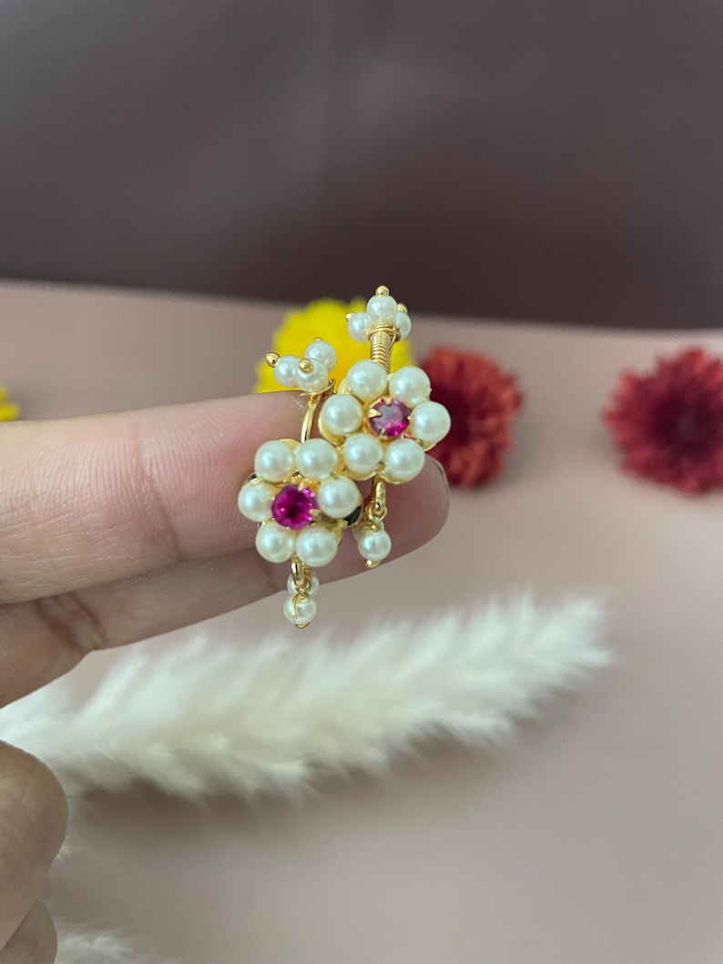Pode incluir: Brincos florais dourados com detalhes de p&eacute;rolas brancas e uma gema rosa central. Os brincos t&ecirc;m um design delicado com pequenas p&eacute;rolas penduradas. O fundo &eacute; rosa suave com flores desfocadas.