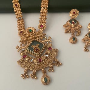 May include: Gold-toned necklace and earring set. The necklace features a central pendant with a green square design and floral accents, complemented by red and white gemstones. The earrings mirror the pendant's design, with a teardrop-shaped green stone at the bottom.