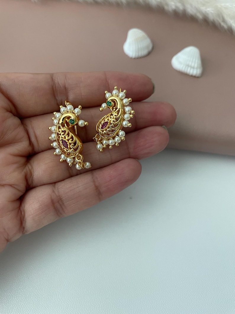 Pode incluir: Brincos dourados em forma de paisley com detalhes intrincados em filigrana. Cada brinco apresenta uma pedra preciosa vermelha central e uma borda circundante de pequenas p&eacute;rolas brancas. Uma pequena pedra preciosa verde tamb&eacute;m &eacute; vis&iacute;vel. Os brincos s&atilde;o exibidos em uma m&atilde;o.