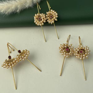 May include: Gold-toned earrings with a floral design. Each earring features a cluster of small, white pearls surrounding a central, round, red gemstone. The earrings have a long, straight post and a curved hook.