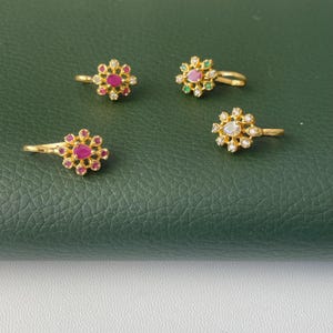 May include: Four gold-colored nose rings with floral designs. Each ring features a central gemstone surrounded by smaller stones. The gemstones are red, pink, green, and clear. The rings are displayed on a dark green surface.