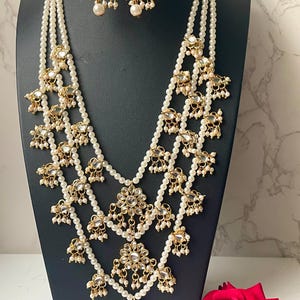May include: A four-strand pearl necklace with gold accents and a large pendant. The necklace has multiple layers of pearls and gold embellishments, including small floral designs and dangling pearls. The pendant is a large, ornate floral design with a mirrored center.