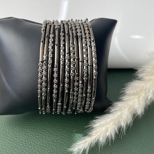 Op de afbeelding: Een set donkergrijze armbanden met ingewikkelde bloemen- en kralenversieringen. De armbanden worden gepresenteerd op een zwarte standaard tegen een groene achtergrond, met een takje witte, veervormige plant.