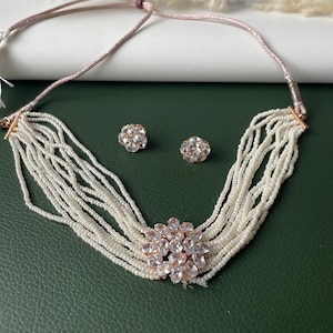 May include: A necklace and earring set featuring a multi-strand pearl necklace with a floral pendant. The pendant and matching earrings are adorned with clear crystals. The necklace has a pink cord.