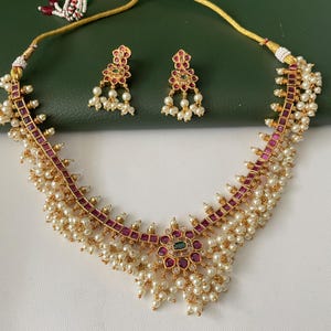 Puede incluir: Un conjunto de collar y pendientes dorados a juego. El collar presenta un diseño floral central con piedras preciosas rojas y verdes, rodeado de pequeñas perlas. El collar y los pendientes tienen una cadena dorada y están sobre un fondo verde.