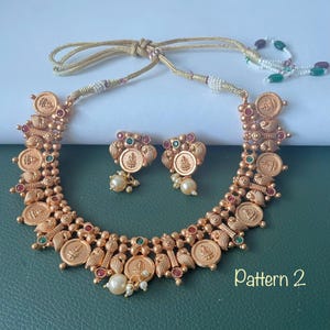 May include: A gold-toned necklace and earring set featuring circular coin-style pendants with embossed figures, accented by small red and green stones and white pearls. The necklace includes a gold cord with white and green bead details. Pattern 2.