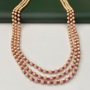 May include: A three-strand necklace featuring gold-colored, textured beads interspersed with small, red beads. The necklace is displayed against a neutral background, highlighting the jewelry's design and color.