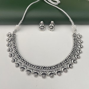 May include: A silver-toned necklace and matching earrings set. The necklace features a beaded design with floral and crescent-shaped accents. The earrings are small, round studs with similar detailing. The jewelry is displayed on a white surface.