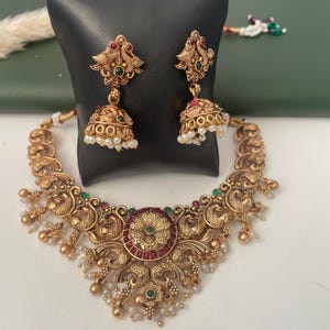 May include: A gold-toned necklace and matching jhumka earring set featuring intricate peacock motifs, embedded with small red and green stones, and accented with dangling white pearl beads. The metalwork displays a textured, antique finish.