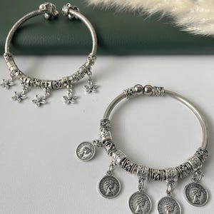 Könnte beinhalten: Zwei silberfarbene Armreifen. Einer hat baumelnde Blumenanhänger, der andere münzförmige Anhänger. Beide haben dekorative, perlenbesetzte Abschnitte. Die Armreifen sind vor einem weißen und grünen Hintergrund zu sehen.