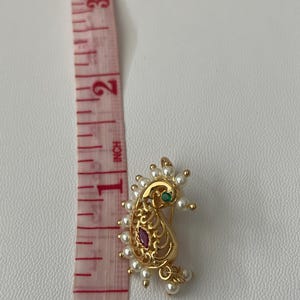 Pode incluir: Anel de nariz dourado em forma de paisley com um design de filigrana, adornado com uma pedra preciosa vermelha e verde, e rodeado por pequenas p&eacute;rolas brancas. O anel de nariz tem aproximadamente 2,5 cm de comprimento.