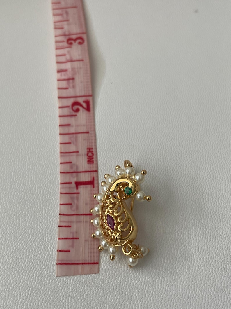 Pode incluir: Anel de nariz dourado em forma de paisley com um design de filigrana, adornado com uma pedra preciosa vermelha e verde, e rodeado por pequenas p&eacute;rolas brancas. O anel de nariz tem aproximadamente 2,5 cm de comprimento.