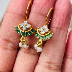 Pode incluir: Brincos dourados com um design floral. Cada brinco apresenta um grupo de pedras preciosas brancas no centro, rodeado por um anel de pequenas pedras verdes. Pequenas p&eacute;rolas brancas pendem da parte inferior de cada brinco.