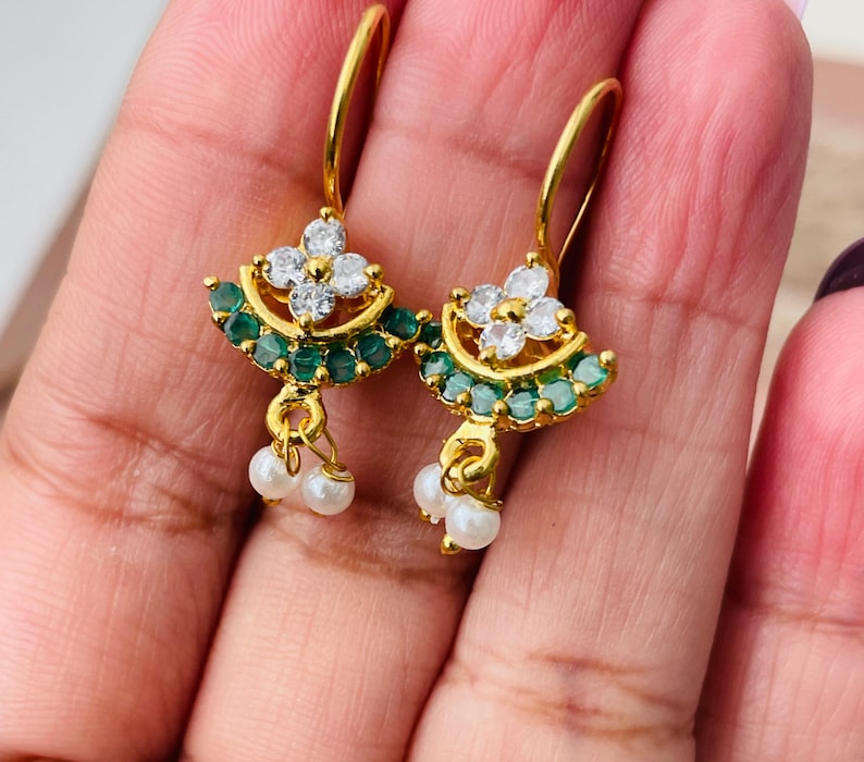 Pode incluir: Brincos dourados com um design floral. Cada brinco apresenta um grupo de pedras preciosas brancas no centro, rodeado por um anel de pequenas pedras verdes. Pequenas p&eacute;rolas brancas pendem da parte inferior de cada brinco.