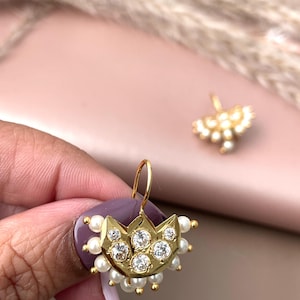 Pode incluir: Brincos dourados com um design semelhante a uma coroa, com pedras preciosas transparentes e pequenas p&eacute;rolas brancas. Os brincos t&ecirc;m um fecho de gancho. A imagem mostra um close-up do brinco, com um segundo brinco no fundo.
