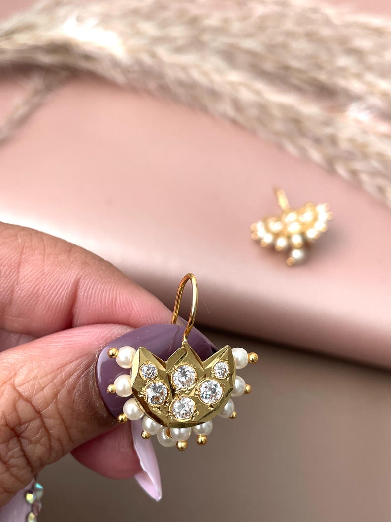 Pode incluir: Brincos dourados com um design semelhante a uma coroa, com pedras preciosas transparentes e pequenas p&eacute;rolas brancas. Os brincos t&ecirc;m um fecho de gancho. A imagem mostra um close-up do brinco, com um segundo brinco no fundo.