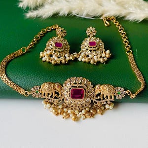 May include: A gold-toned necklace and earring set. The necklace features a central rectangular ruby-colored stone, surrounded by smaller clear stones and elephant motifs. The earrings mirror the design with ruby-colored stones and pearl accents. The set is on a green surface.