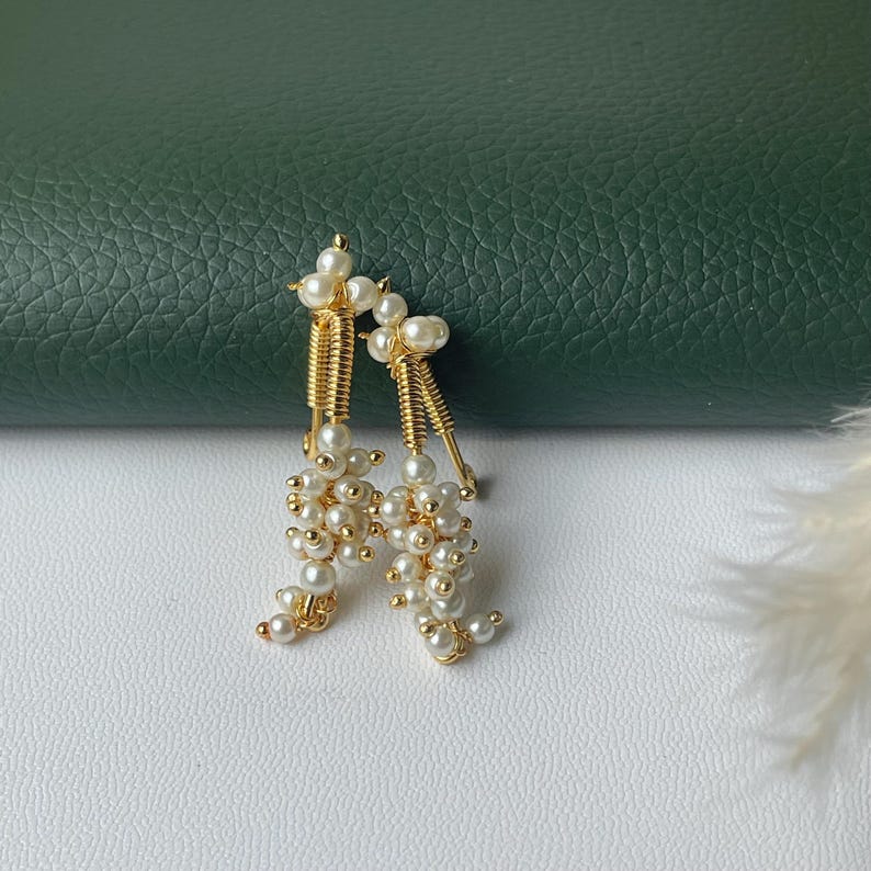 Pode incluir: Um par de brincos dourados com aglomerados de pequenas p&eacute;rolas brancas. Os brincos t&ecirc;m um design &uacute;nico com um elemento vertical texturizado e uma cascata de p&eacute;rolas, criando um visual delicado e elegante. Os brincos est&atilde;o sobre uma superf&iacute;cie branca.