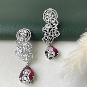 May include: A pair of silver-toned drop earrings. Each earring features a cluster of clear, sparkling stones in an openwork design, with a large, teardrop-shaped, ruby-red stone at the bottom. The earrings are displayed against a green background.