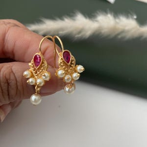 Pode incluir: Brincos dourados com uma pedra vermelha em forma de gota e um grupo de pequenas p&eacute;rolas brancas. Os brincos t&ecirc;m um design de filigrana decorativo e um fecho de gancho.