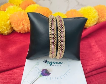 May include: Two gold bangles with a delicate, intricate design and pink stones. The bangles are displayed on a black cushion and a white box with the text "nysaa... JEWELRY" and "Etsy: @nysaaajewelry".