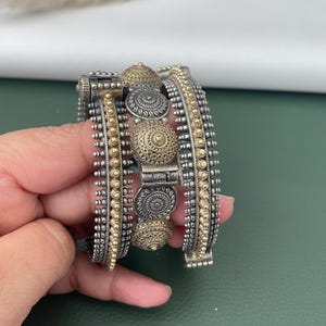 May include: A silver and gold-toned cuff bracelet with intricate detailing. The bracelet features a series of decorative elements, including circular patterns and beaded accents. The design incorporates both silver and gold tones, creating a striking contrast.