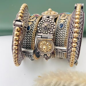 May include: A set of three hinged metal bangles featuring a mix of silver and gold tones. The bracelets display intricate filigree patterns, beaded edges, and small embedded blue and red stones, with central floral and circular embossed motifs.