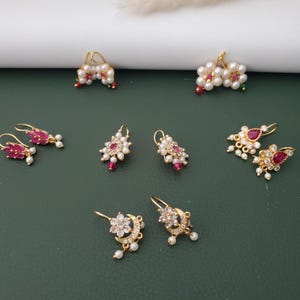 May include: A collection of gold-tone earrings featuring various designs, including floral motifs with white pearls and pink stones, crescent moon shapes with clear crystals, and teardrop-shaped red stones accented with small white pearls and clear crystals.