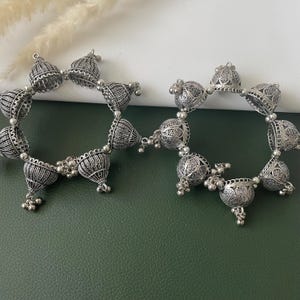 May include: Two silver-toned, ornate bangles. Each bangle features a series of bell-shaped charms with intricate detailing and small dangling accents. The bangles are displayed on a green and white surface.