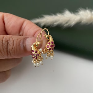 Pode incluir: Brincos dourados com design de pav&atilde;o e detalhes em vermelho e verde. Os brincos apresentam um design de pav&atilde;o com pedras preciosas vermelhas e pequenos pingentes de p&eacute;rolas brancas. Os brincos s&atilde;o segurados por uma m&atilde;o.