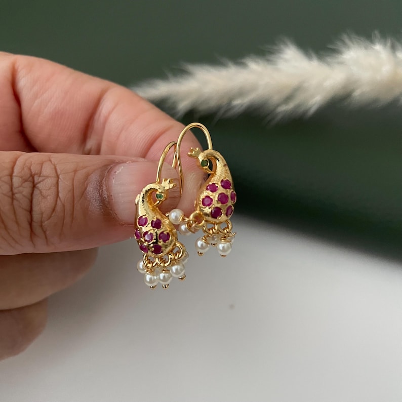 Pode incluir: Brincos dourados com design de pav&atilde;o e detalhes em vermelho e verde. Os brincos apresentam um design de pav&atilde;o com pedras preciosas vermelhas e pequenos pingentes de p&eacute;rolas brancas. Os brincos s&atilde;o segurados por uma m&atilde;o.