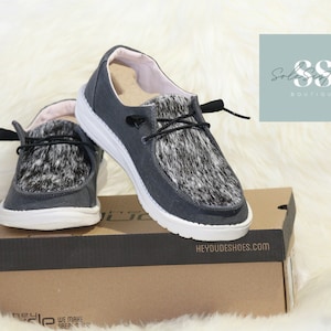 May include: A pair of gray canvas slip-on shoes with a white sole and black and white patterned accents. The shoes are displayed on a cardboard box with the text "HEYDUDESHOES.COM". The shoes are from the Solesisters Boutique.