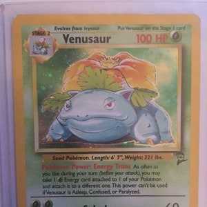 May include: A blue and green Pokemon trading card featuring Venusaur, a Grass-type Pokemon. The card has a light blue background with a green border. The card shows Venusaur with a large flower on its head. The card text reads "Venusaur, Seed Pokemon, Length: 6' 7", Weight: 221 lbs. Pokemon Power: Energy Trans. Solarbeam 60. This plant blooms when it is absorbing solar energy. It stays on the move to seek sunlight. LV. 67 #3. 18/130*"