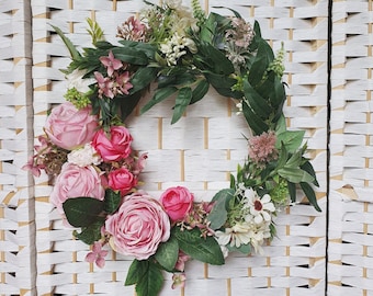 May include: A pink and white floral wreath with green leaves. The wreath is made of artificial flowers and is hanging on a white wicker wall.
