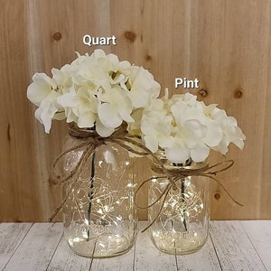 May include: Two mason jars, one quart and one pint, filled with white hydrangeas and fairy lights. The jars are tied with twine and placed on a wooden surface.