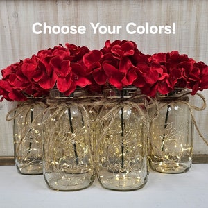 May include: Six clear glass jars with string lights inside, decorated with red hydrangea flowers. The jars are arranged in a row on a wooden surface. The text "Choose Your Colors!" is written above the jars.