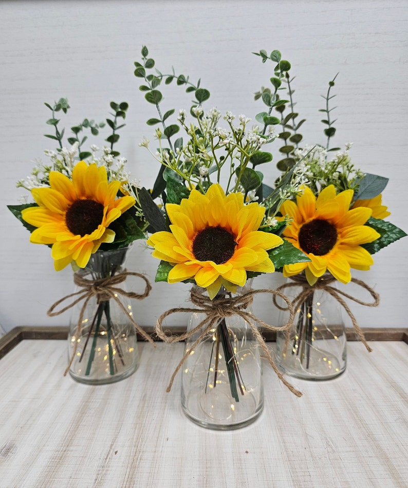 May include: Three clear glass vases with string tied around the top, each vase contains a bouquet of yellow sunflowers, white baby's breath, and green eucalyptus leaves. The vases are lit with white fairy lights.