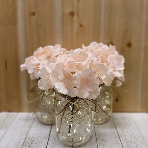 Puede incluir: Tres tarros de cristal con luces en su interior, decorados con cuerda y llenos de flores de hortensia rosas.