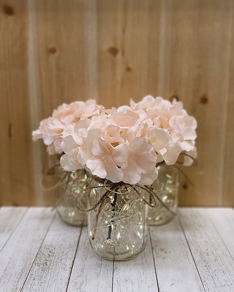 Puede incluir: Tres tarros de cristal con luces en su interior, decorados con cuerda y llenos de flores de hortensia rosas.