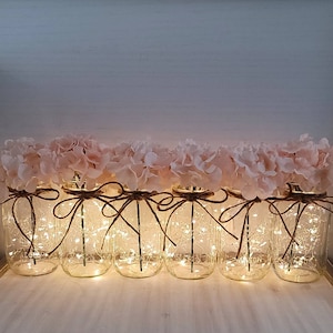 Puede incluir: Una fila de tarros de cristal decorados con hortensias rosas y envueltos en cuerda. Cada tarro contiene una cadena de luces de hadas, creando un ambiente c&aacute;lido y acogedor.