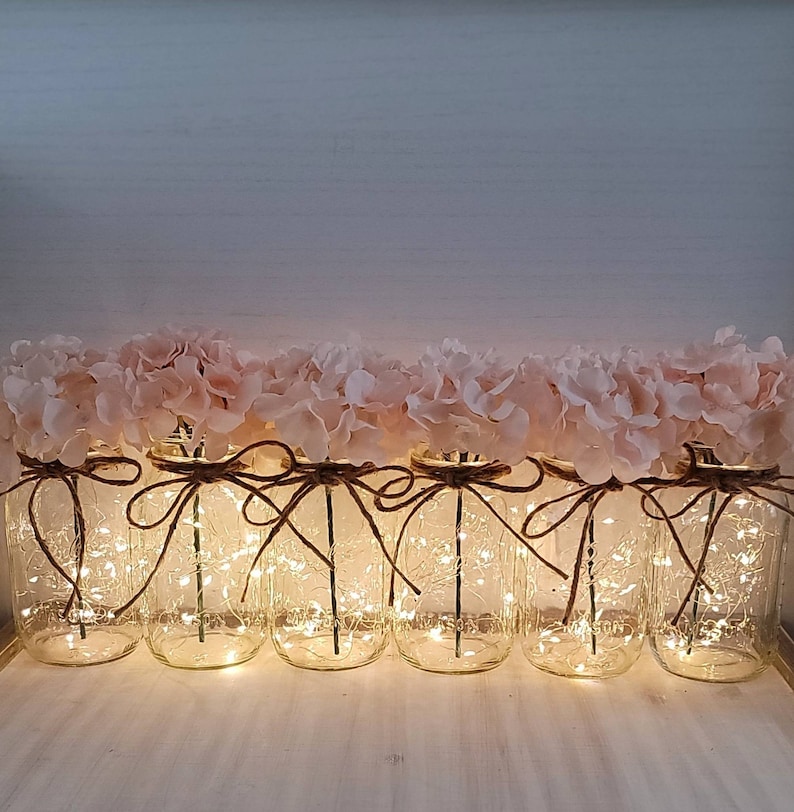 Puede incluir: Una fila de tarros de cristal decorados con hortensias rosas y envueltos en cuerda. Cada tarro contiene una cadena de luces de hadas, creando un ambiente c&aacute;lido y acogedor.