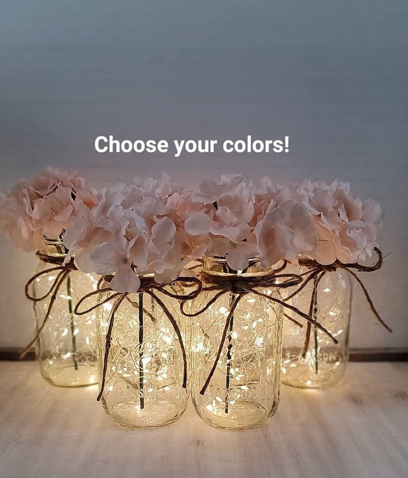 Puede incluir: Cinco tarros de cristal con luces de hadas en el interior y flores de hortensia rosa en la parte superior. Los tarros est&aacute;n atados con cuerda y tienen un aspecto r&uacute;stico. &iexcl;Elige tus colores!