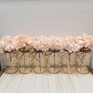 Puede incluir: Seis tarros de cristal con luces de cadena y flores de hortensia rosas. Los tarros est&aacute;n atados con cuerda y tienen un aspecto r&uacute;stico.