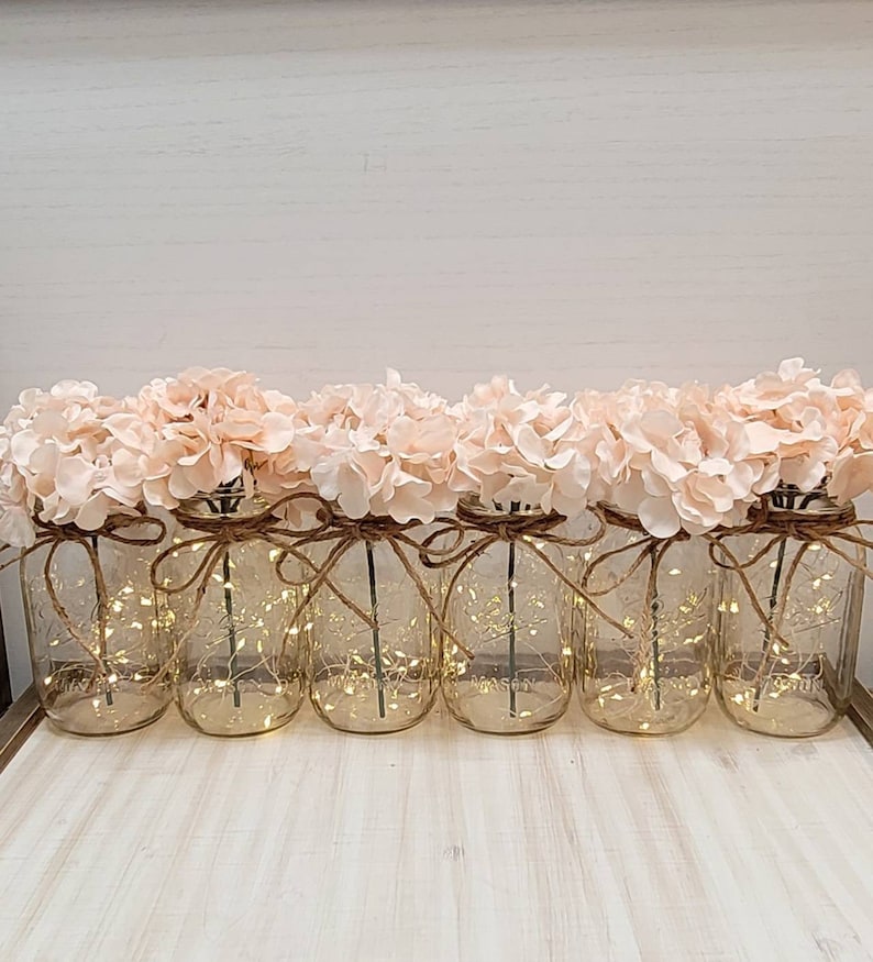 Puede incluir: Seis tarros de cristal con luces de cadena y flores de hortensia rosas. Los tarros est&aacute;n atados con cuerda y tienen un aspecto r&uacute;stico.