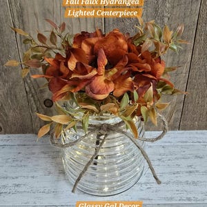 K&ouml;nnte beinhalten: Ein herbstliches, beleuchtetes Kunst-Hortensien-Gesteck mit rostfarbenen Seidenblumen und gr&uuml;nem Laub in einem klaren, horizontal gerippten Glasgef&auml;&szlig;. Das Glas ist mit einer nat&uuml;rlichen Jutekordel verziert. Text: Fall Faux Hydrangea Lighted Centerpiece.