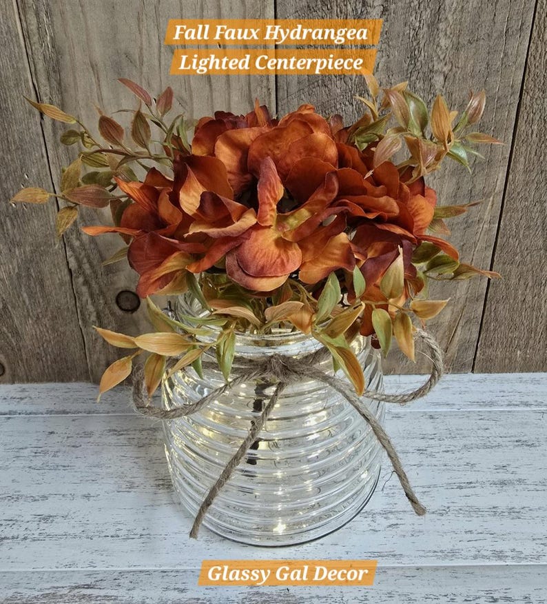 K&ouml;nnte beinhalten: Ein herbstliches, beleuchtetes Kunst-Hortensien-Gesteck mit rostfarbenen Seidenblumen und gr&uuml;nem Laub in einem klaren, horizontal gerippten Glasgef&auml;&szlig;. Das Glas ist mit einer nat&uuml;rlichen Jutekordel verziert. Text: Fall Faux Hydrangea Lighted Centerpiece.