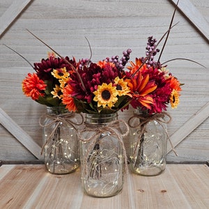 May include: Three glass mason jars with autumn-colored floral arrangements. Each jar is filled with string lights and tied with twine. The flowers include sunflowers, dahlias, and berries in shades of orange, yellow, and burgundy.
