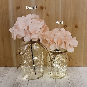 Puede incluir: Dos tarros de cristal, uno etiquetado como "cuarto" y el otro como "pinta", est&aacute;n llenos de hortensias rosas y luces de hadas. Los tarros est&aacute;n atados con cuerda.
