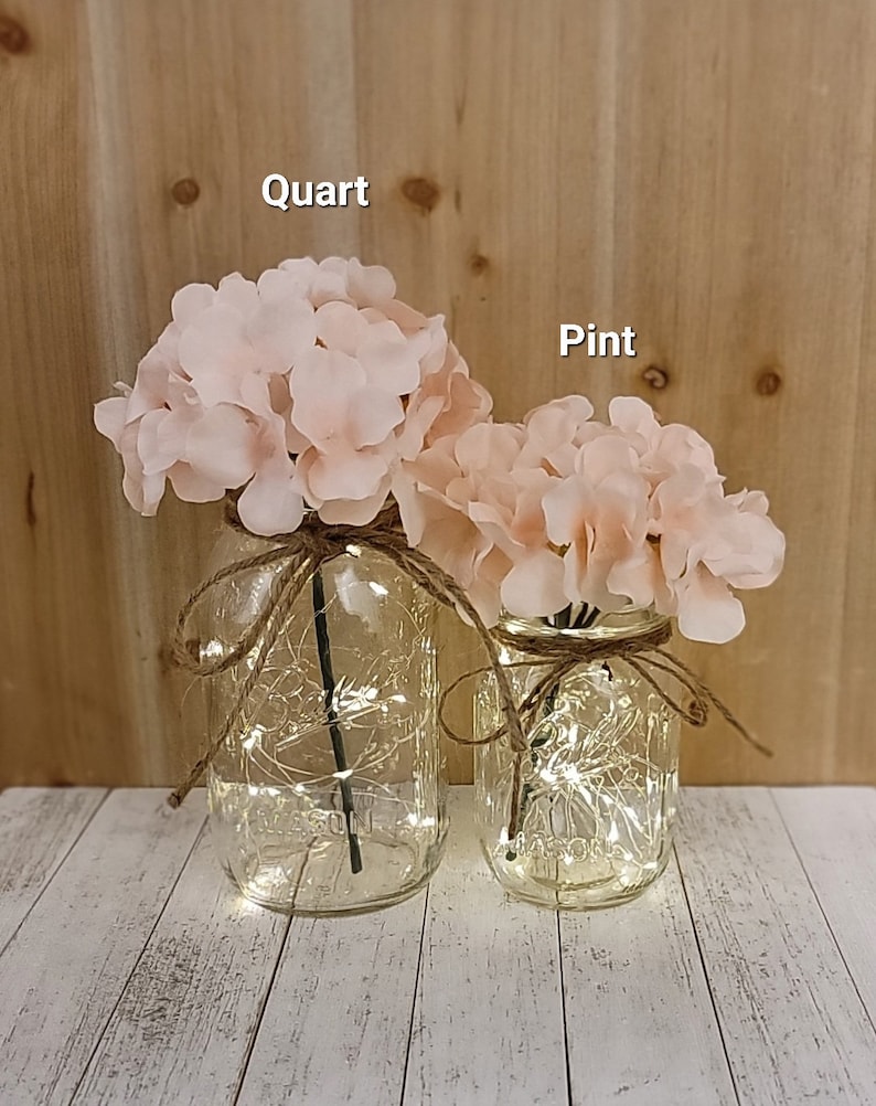 Puede incluir: Dos tarros de cristal, uno etiquetado como "cuarto" y el otro como "pinta", est&aacute;n llenos de hortensias rosas y luces de hadas. Los tarros est&aacute;n atados con cuerda.
